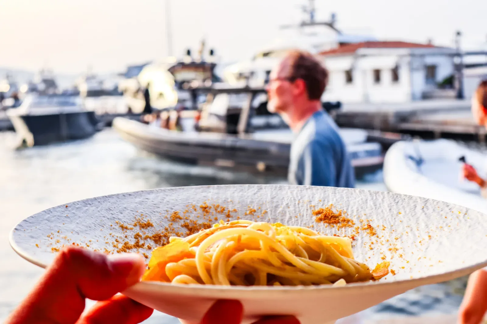 Un piatto di spaghetti alla bottarga tenuto in mano davanti al porto di Porto Rotondo, con i gommoni e gli yacht sfocati alle spalle