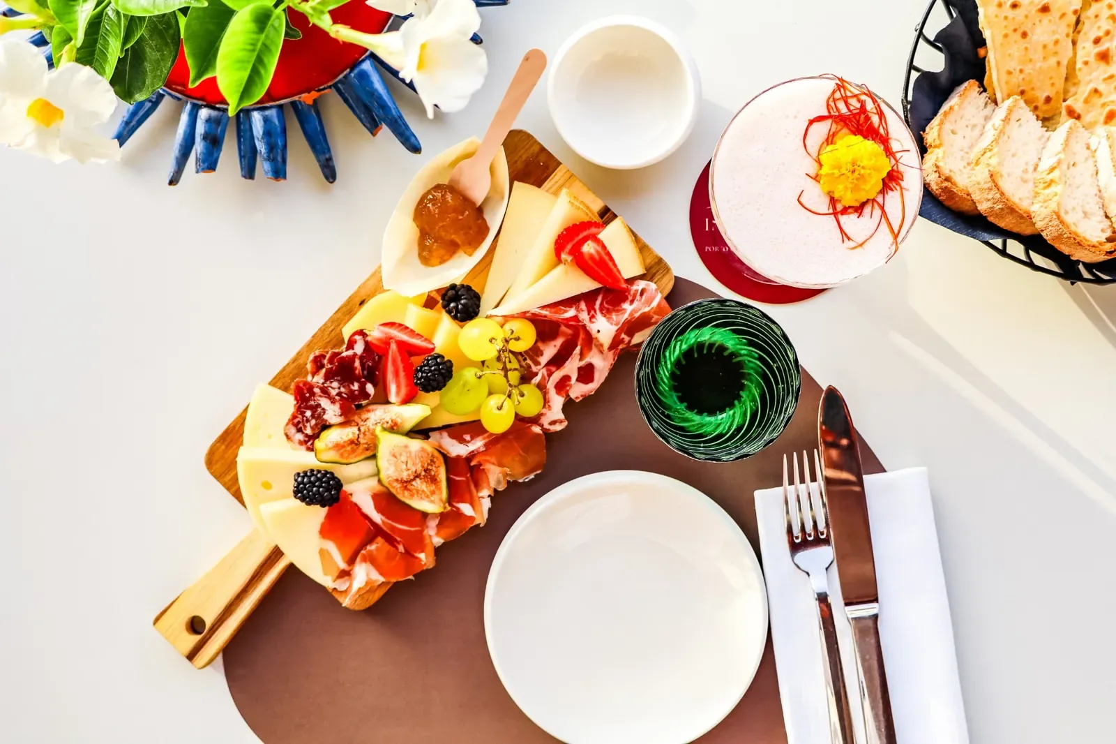 Tagliere di formaggi, prosciutto, fichi e frutti di bosco visto dall'alto, con un cocktail decorato, il pane e la tavola apparecchiata