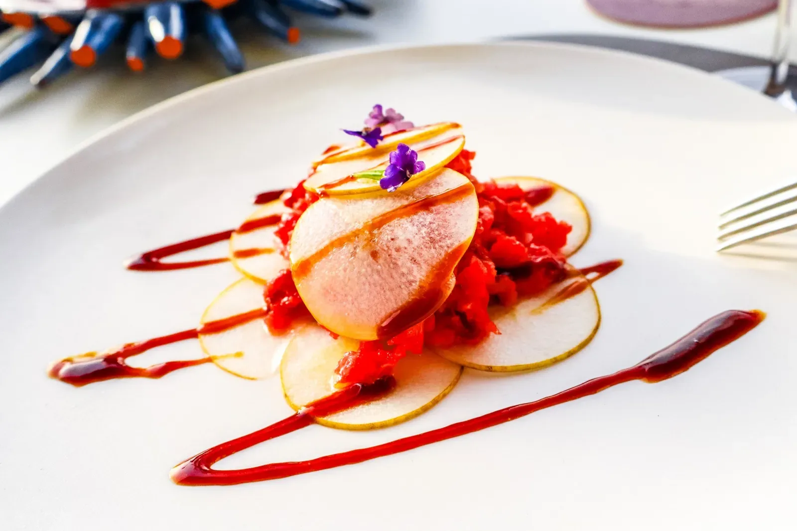 Tartare coperta da fette sottili di pera, con riduzione rossa e fiori di lavanda su un piatto bianco